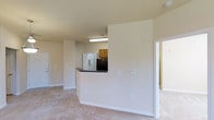This single-floor residential unit presents a practical and well-organized open-concept layout designed for comfortable everyday living. The living area features neutral-toned carpeting that extends throughout, creating a warm and cohesive feel across the space. A defining architectural feature is the half-wall pass-through that separates the kitchen from the living area, providing an open connection between the two spaces while maintaining a sense of structure. The kitchen is equipped with warm wood-toned cabinetry, a dark countertop along the pass-through bar, a refrigerator, and a stacked washer/dryer unit, offering both style and convenience in a compact, efficient footprint. Overhead fluorescent lighting in the kitchen ensures the space is well-lit. The walls throughout are finished in a soft, neutral beige tone, contributing to a bright and airy ambiance. An adjacent room, accessible through a doorway on the right, appears to offer additional living or sleeping space. The overall design emphasizes functionality, with a straightforward floor plan that maximizes usability. The clean, move-in-ready condition of the space makes it an ideal option for those seeking a well-maintained, single-level residential unit.