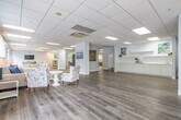 Kings Reserve is a generously sized single-floor community facility thoughtfully designed to accommodate a variety of social and recreational activities. The expansive main gathering area features light wood-look flooring throughout, a drop-panel ceiling with recessed lighting, and an abundance of natural light streaming through floor-to-ceiling windows that frame views of the surrounding mature tree canopy. Multiple round white dining tables with cross-back chairs are arranged throughout this open area, creating an inviting setting for communal dining and socializing. A wall-mounted television and a decorative blue sideboard provide functional and aesthetic focal points. A classic upright piano adds a warm, residential character to the space.

The kitchenette area is outfitted with extensive white shaker-style cabinetry, open shelving, a continuous countertop with an integrated sink, and a commercial-grade coffee station, providing ample support for food and beverage service. An adjacent room features upper and lower white cabinetry with a rich wood-toned countertop and a central dining table, offering a more intimate setting. Large glass panel doors open directly to an outdoor patio furnished with seating, seamlessly connecting the interior to the exterior environment. Colorful artwork displayed throughout adds a vibrant, welcoming ambiance to the entire space.