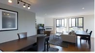 This single-floor space showcases a contemporary open-plan design that seamlessly integrates the dining and living areas into one cohesive environment. The dining area features a dark espresso-toned table paired with matching upholstered chairs, anchored by a track lighting fixture overhead that adds a warm, directional glow. A partial wall creates a subtle visual divide between the dining space and the rest of the room, adding architectural interest without compromising the open feel.

The living area is centered around a sleek wall-mounted electric fireplace with a minimalist linear design, flanked by a flat-screen television on a dark media console. A comfortable grey sectional sofa and coordinating armchairs are arranged around a rectangular coffee table, all grounded by a plush area rug with rich texture. Large floor-to-ceiling windows and a glass sliding door flood the space with natural light and offer views of the surrounding outdoor greenery. The overall ambiance is clean, modern, and refined, with a neutral color palette of whites, greys, and dark wood tones throughout.