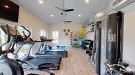 This fitness center offers a comprehensive workout environment designed to support a wide range of exercise routines. The space features vaulted ceilings that lend an open, airy feel, complemented by recessed lighting and a ceiling fan that help maintain a comfortable atmosphere during workouts. Light wood-style flooring runs throughout, providing a clean and functional surface underfoot.

The cardio section is well-appointed with multiple TRUE-brand treadmills, an elliptical trainer, and a rowing machine, all positioned to take advantage of the natural light streaming in through large windows and glass sliding doors that offer views of the surrounding greenery. A wall-mounted television is available for entertainment during cardio sessions.

The strength training area includes an Apollo multi-station cable machine, a dedicated leg extension/curl bench, and a full rack of dumbbells in varying weights. Functional training is supported by a freestanding rack stocked with resistance bands, medicine balls, and stability balls in multiple sizes. Exercise mats are also available for floor work and stretching.

Motivational framed prints line the walls, adding an encouraging atmosphere throughout. A water fountain station near the entrance provides added convenience. The abundant natural light, vaulted ceilings, and diverse equipment selection make this a thorough and inviting fitness facility.