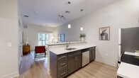Located at 69 Marlborough Street in Portland, Connecticut, this thoughtfully designed one-bedroom, one-bathroom residence offers a efficient and modern living experience. The open-concept kitchen and living area serves as the heart of the home, featuring light hardwood-style flooring, neutral toned walls, and an abundance of natural light. The kitchen is outfitted with dark espresso cabinetry, white quartz countertops, stainless steel appliances including a full-size refrigerator, microwave, and dishwasher, and a peninsula with bar seating for three — ideal for casual dining and entertaining. Pendant lighting and a track light fixture illuminate the space stylishly. The living area opens directly onto a private balcony through a sliding glass door, offering views of the surrounding community courtyard. The bedroom is a comfortable retreat featuring a large window, warm hardwood-style flooring, and a closet. The full bathroom mirrors the kitchen's design language with dark wood cabinetry, a white quartz vanity top, a large mirror, and a tub-shower combination with clean white subway tile. The residence is part of a modern multi-story community with an expansive paved courtyard and contemporary architectural detailing.