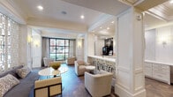 This clubroom in Houston, TX presents a refined, single-level communal space designed with upscale finishes and thoughtful architectural detailing. The layout is open and expansive, organized into distinct yet flowing zones that encourage both socializing and relaxation.

The lounge areas are furnished with plush gray and taupe seating arrangements, including sofas, armchairs, and accent tables, anchored by a neutral-toned area rug. A striking large-scale abstract artwork serves as a focal point in one seating zone, flanked by antique mirrored panels that amplify natural light and add depth to the space. Coffered ceilings with crown molding and recessed lighting lend an air of classical elegance throughout.

A standout decorative screen wall, featuring a geometric gold-and-white lattice pattern, elegantly divides portions of the space while maintaining visual connectivity. Large steel-framed windows flood the interior with natural light and frame views of the surrounding streetscape.

The wet bar area is well-appointed with white cabinetry, marble countertops, a stainless steel sink, a mounted flat-screen television, and textured tile backsplash. A coffee station and small appliances make the space fully functional for resident gatherings.

Underfoot, a richly patterned herringbone wood-look tile floor unifies the entire space, adding warmth and visual sophistication to this polished communal retreat.