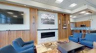 This single-floor space offers a thoughtfully designed interior that blends warmth and professionalism. The centerpiece of the lounge area is a sleek, linear stone fireplace framed by rich wood-paneled cabinetry, creating a refined and inviting focal point. Flanking the fireplace are elegant wall sconces that cast a soft, ambient glow, while recessed ceiling lighting ensures the space is well-lit throughout. Built-in shelving and cabinetry on either side of the fireplace provide functional display and storage, adding to the polished aesthetic. A large framed cityscape photograph mounted above the fireplace adds a local character and visual interest to the room. Comfortable blue accent chairs are arranged around a contemporary coffee table atop a multicolored area rug, offering a casual seating arrangement suited for conversation or relaxation. The drop ceiling with recessed lighting maintains a clean, modern appearance. Visible in the background is a kitchenette or refreshment area featuring a tiled backsplash and stainless steel appliances, adding convenience to the space. Overall, the interior strikes a balance between a professional commercial environment and a warm, hospitable atmosphere.