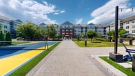 Located at 8 Norden Pl in Norwalk, Connecticut, this thoughtfully designed outdoor amenity space serves as a vibrant community hub set within a modern multi-story residential complex. The grounds feature a striking combination of brick and siding architecture that frames the courtyard on multiple sides, creating an enclosed yet open-air atmosphere.

At the heart of the space is a generously sized resort-style swimming pool with a stamped concrete surround, outfitted with lounge chairs and shaded umbrellas in bold blue and red tones. Adjacent to the pool, a colorful pickleball court finished in blue and yellow adds a recreational focal point, bordered by lush artificial turf and mature landscaping.

A spacious patio area provides multiple seating arrangements, including Adirondack chairs, bistro tables, and a prominent stone fire pit feature — all strung with overhead café lighting for evening ambiance. A dedicated lawn game area with a ping-pong table and additional outdoor furniture offers further recreational options.

Landscaping throughout includes manicured greenery, ornamental trees, trimmed hedgerows, and flowering plants that soften the hardscape. Wide stone-paved walkways connect each zone seamlessly, while a gated entry provides secure access from the surrounding parking area. The space is bathed in natural light under open skies, offering a welcoming and active outdoor environment.
