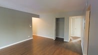 This single-floor space offers a clean, functional layout designed for comfortable everyday living. The main living area is finished with warm-toned hardwood flooring that extends throughout, providing a cohesive and inviting foundation. Walls are painted in a neutral grey tone, creating a versatile backdrop suited to a variety of interior styles.

A built-in closet with a standard hanging rod is conveniently integrated into the main room, offering practical storage without occupying additional floor space. White baseboards provide a crisp contrast against both the grey walls and hardwood floors, adding a subtle architectural detail that defines the room's boundaries.

An open doorway visible to the right connects the main space to an adjacent room, suggesting a logical and accessible flow between areas. The layout appears well-proportioned, with sufficient open floor space to accommodate multiple furniture configurations. Overhead lighting contributes to a bright interior atmosphere. Overall, the space presents a straightforward, move-in-ready environment with a neutral palette that allows for personalization.