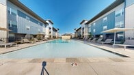 This outdoor amenity space centers around a generously sized resort-style swimming pool, designed to serve as a premier gathering and relaxation destination within a modern residential community. The pool itself is a standout feature — crystal-clear water fills a large rectangular basin with accessible entry points via stainless steel handrails and steps, with a maximum depth of four feet throughout.

The surrounding deck is finished in light-toned concrete, providing ample space for lounging and socializing. Rows of contemporary sling-style chaise lounges with sleek metal frames are arranged along both sides of the pool, accompanied by shade umbrellas that offer relief on sunny days. The open layout encourages both active recreation and leisurely relaxation under wide, open skies.

The pool courtyard is flanked on multiple sides by multi-story residential buildings, clad in a modern combination of vertical board-and-batten siding in cool grey tones and classic light brick masonry. Many of the upper-level units feature private balconies overlooking the pool area. Ornamental iron fencing defines the perimeter of the amenity space, with manicured landscaping including young cypress trees adding greenery and privacy.

A separate amenity building — with large windows and a covered patio — is visible in the background, suggesting additional community facilities nearby. The overall ambiance is clean, contemporary, and resort-inspired.