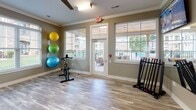 Located at 159 W Kinderton Way in Bermuda Run, North Carolina, this single-floor fitness studio is a well-appointed exercise space designed to support a variety of workout routines. The room features light wood-look flooring throughout, complemented by warm taupe walls adorned with crisp white crown molding, creating a clean and inviting atmosphere.

The studio is equipped with stationary cycling bikes positioned for easy use, alongside a dedicated rack of color-coded dumbbells and a wall-mounted storage rack holding multiple exercise and medicine balls in vibrant colors. A large wall mirror on one side of the room enhances the sense of space and allows users to monitor their form during workouts. A wall-mounted flat-screen television is positioned for visibility throughout the room, adding an entertainment element to exercise sessions.

Natural light fills the studio through multiple large windows fitted with white blinds, offering views of the surrounding community grounds. A glass door with transom windows provides direct access to an outdoor covered patio area, which is furnished with seating and overlooks what appears to be a pool area, seamlessly connecting indoor fitness with outdoor amenities. The space is finished with ceiling fans and recessed lighting to ensure comfort during workouts. A branded frosted glass panel adds a polished, professional touch to the overall design.