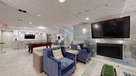 Located at 163 Franklin St in Stamford, Connecticut, this elegantly designed amenity lounge offers a well-appointed gathering space for socializing and recreation. The open floor plan is unified by light-toned herringbone-patterned flooring that runs throughout, creating a bright and cohesive aesthetic.

The lounge is divided into distinct functional zones. A central game area features a full-size billiards table as its focal point, providing ample room for play. Adjacent to it, a wet bar with a curved arched ceiling is outfitted with a long white countertop, bar stool seating, pendant lighting, and a wall-mounted television — ideal for entertaining.

A dedicated lounge seating area showcases plush blue velvet armchairs and a neutral sofa arranged around a fireplace set into a striking floor-to-ceiling mosaic tile accent wall. A large flat-screen television is mounted above the fireplace, making this zone a natural media hub. Wall sconces on either side of the fireplace wall add warm, ambient lighting.

A sculptural glass-and-steel spiral staircase serves as a dramatic architectural centerpiece, connecting this level to the floor above. The space also includes elevator access, recessed ceiling lighting, and additional seating alcoves. The overall design palette of white, silver, and navy blue delivers a modern, upscale ambiance throughout.