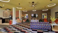 This single-floor space makes an immediate impression with its bold, retro-inspired aesthetic and carefully curated design elements. The exposed brick accent wall serves as a striking architectural centerpiece, anchoring the room and adding warmth and texture to the overall environment. A large flat-screen television is mounted prominently on the brick wall, making the space well-suited for communal viewing and social gatherings.

The interior is illuminated by a combination of circular pendant ring lights and a ceiling fan, which together create a layered lighting effect that is both functional and visually dynamic. The yellow-toned ceiling adds a warm, energetic contrast to the rich brick tones below.

Furnishings are deliberately eclectic and colorful — a vibrant patterned sofa in multicolored hues takes center stage, complemented by sleek orange accent chairs that reinforce the retro mid-century modern vibe. Abstract artworks adorn the walls, adding an artistic and creative dimension to the space.

The flooring features a bold, multicolored geometric carpet that ties the design elements together and adds to the playful energy of the room. Lush potted plants provide a touch of greenery, softening the bold palette and bringing a sense of life and freshness. Overall, this space exudes a dynamic, creative, and inviting character ideal for socializing and collaboration.