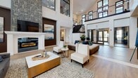 Located at 161 Grande Oaks Dr in Charleston, SC, this community clubhouse at Grande Oaks Place offers an impressive and thoughtfully designed communal space. The centerpiece of the lobby is a dramatic double-height ceiling that creates an airy, grand atmosphere, complemented by an open mezzanine level with sleek metal railings visible above. A striking floor-to-ceiling stone fireplace anchors one side of the space, flanked by curated artwork that adds a sophisticated touch to the interior. Light hardwood flooring flows throughout, unifying the open layout. The seating area is furnished with contemporary white upholstered chairs, plush sofas with navy accent pillows, and gold-accented coffee tables, creating an inviting and stylish environment. Large windows and glass entry doors allow abundant natural light to fill the space, with views to the outdoor surroundings. Lush indoor plants in sleek black planters add warmth and a natural element to the modern design palette. A separate office or leasing area, accessible through glass-paned French doors, is visible from the main lobby and features the Grande Oaks branding. The overall ambiance is polished and welcoming, blending contemporary design with comfortable, functional spaces suited for both social gatherings and day-to-day community needs.