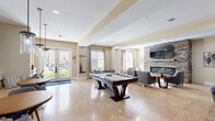 Located at 1600 Granby Street in Norfolk, Virginia, this well-appointed community lounge space offers a thoughtfully designed single-floor layout that blends comfort and functionality. The open-concept interior is anchored by warm neutral tones, travertine tile flooring, and recessed lighting throughout, creating a bright and welcoming atmosphere.

The main gathering area serves as the centerpiece of the space, featuring a full-size pool table, stylish leather seating, and pendant lighting that adds a sophisticated touch. Large windows and glass doors flood the room with natural light while offering views to the outside. A cozy lounge nook showcases a stunning floor-to-ceiling stacked stone fireplace with a mounted flat-screen television, framed by upholstered chairs and a classic round wooden side table — ideal for relaxed conversation.

Multiple dining areas with round tables and modern chairs are positioned throughout, while a mirrored wall accent enhances the sense of openness. A glass-enclosed area visible from the main lounge provides additional workspace or meeting space. The kitchen features grey shaker-style cabinetry with gold hardware, granite countertops, a subway tile backsplash, stainless steel appliances, and a double sink — perfect for hosting and entertaining. A striking stacked stone column connects the kitchen to the lounge, reinforcing the cohesive design aesthetic.