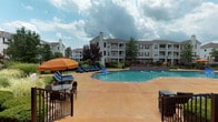 Located at 1000 Applerock Dr in O'Fallon, Missouri, this outdoor pool area serves as a central amenity within the residential community. The space is anchored by a generously sized swimming pool with clear blue water, accented by a water feature along one edge. The surrounding stamped concrete deck provides ample room for outdoor lounging, furnished with blue chaise lounge chairs and a shaded seating area complete with an orange market umbrella and bistro-style table and chairs — ideal for relaxing on sunny days.

Lush landscaping borders the pool deck, incorporating ornamental grasses, shrubs, and mature trees that add both privacy and greenery to the environment. A charming covered porch runs along the adjacent building facade, constructed with classic brick detailing and white-trimmed, multi-pane windows. The porch is lined with traditional white rocking chairs, offering a welcoming spot to unwind and take in views of the pool area. Entry to the adjacent interior space is provided through a bold red-framed glass door.

The multi-story residential buildings surrounding the pool feature private balconies, traditional rooflines with chimneys, and well-maintained exteriors, contributing to a cohesive and inviting community atmosphere. The open-sky setting allows for abundant natural light throughout the day, making this pool area a standout outdoor retreat within the O'Fallon community.