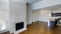 This single-floor space showcases a thoughtfully designed open layout that seamlessly connects the living and dining areas. The living room serves as the focal point of the space, anchored by a classic fireplace with a white-painted mantle and marble tile surround, adding both warmth and visual interest to the room. Rich, dark hardwood flooring runs throughout, providing a warm contrast against the neutral, light-toned walls.

Elevated ceilings lend a sense of openness and grandeur to the living area, further enhanced by carefully detailed crown molding that frames the upper walls. The transition into the adjoining dining area is defined by a subtle architectural divide, where a decorative chandelier hangs overhead, providing an elegant lighting focal point. Crown molding continues throughout, maintaining a cohesive design language across both spaces.

Natural light filters into the space, complementing the warm tones of the hardwood floors and neutral palette. A hallway visible from the main living area provides access to additional rooms, suggesting a well-organized and functional floor plan. Overall, the space offers a classic, understated aesthetic with refined architectural details throughout.