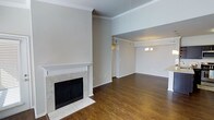This single-floor space showcases a thoughtfully designed open layout that seamlessly connects the living and dining areas. The living room serves as the focal point of the space, anchored by a classic fireplace with a white-painted mantle and marble tile surround, adding both warmth and visual interest to the room. Rich, dark hardwood flooring runs throughout, providing a warm contrast against the neutral, light-toned walls.

Elevated ceilings lend a sense of openness and grandeur to the living area, further enhanced by carefully detailed crown molding that frames the upper walls. The transition into the adjoining dining area is defined by a subtle architectural divide, where a decorative chandelier hangs overhead, providing an elegant lighting focal point. Crown molding continues throughout, maintaining a cohesive design language across both spaces.

Natural light filters into the space, complementing the warm tones of the hardwood floors and neutral palette. A hallway visible from the main living area provides access to additional rooms, suggesting a well-organized and functional floor plan. Overall, the space offers a classic, understated aesthetic with refined architectural details throughout.