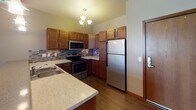 Located on Calico Drive South in Fargo, North Dakota, this single-floor unit offers a well-organized layout designed for comfortable everyday living. The open-concept living and dining area flows naturally into the kitchen, which is appointed with warm-toned wood cabinetry, a mosaic tile backsplash, stainless steel appliances including a refrigerator, electric range, and built-in microwave, and a peninsula with a raised breakfast bar illuminated by pendant lighting. Rich hardwood-style flooring runs throughout the kitchen and dining space, while plush carpet adds warmth to the living area and bedrooms. The living room features recessed lighting and a sliding glass door fitted with vertical blinds, providing access to an outdoor area and allowing natural light to fill the space. The bedrooms are carpeted and include ceiling fans, windows with exterior views of the surrounding neighborhood, and ample wall space. The bathroom is well-appointed with rich wood cabinetry, a granite-topped vanity, tile flooring, and a full tub-shower combination. Throughout the unit, consistent wood trim accents complement the neutral color palette, creating a cohesive and inviting atmosphere suited to a range of lifestyles.