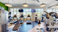 Located at 2130 Leather Fern Dr in Odessa, FL, this modern fitness center is designed to deliver a comprehensive workout experience across a single, thoughtfully organized floor. The space is divided into distinct zones, each purpose-built to serve different training needs. The cardio area is lined with floor-to-ceiling windows that flood the room with natural light and offer sweeping views of the surrounding palm-lined landscape, creating an energizing atmosphere. Multiple treadmills and elliptical machines are positioned to take full advantage of these outdoor vistas, with wall-mounted TVs overhead for entertainment. Adjacent to the cardio zone, a robust strength training area features a wide selection of cable machines, selectorized equipment, a full dumbbell rack, and adjustable benches, all set against large mirrored walls and motivational wall graphics. Pendant lighting and ceiling fans maintain a comfortable environment throughout. A separate, glass-partitioned studio provides dedicated space for spinning bikes and floor exercises, complete with light wood flooring and ample natural light. The overall design blends clean, modern aesthetics with vibrant accents, delivering a welcoming and functional environment for a full range of fitness activities.