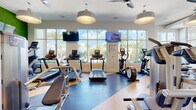 Located at 2130 Leather Fern Dr in Odessa, FL, this modern fitness center is designed to deliver a comprehensive workout experience across a single, thoughtfully organized floor. The space is divided into distinct zones, each purpose-built to serve different training needs. The cardio area is lined with floor-to-ceiling windows that flood the room with natural light and offer sweeping views of the surrounding palm-lined landscape, creating an energizing atmosphere. Multiple treadmills and elliptical machines are positioned to take full advantage of these outdoor vistas, with wall-mounted TVs overhead for entertainment. Adjacent to the cardio zone, a robust strength training area features a wide selection of cable machines, selectorized equipment, a full dumbbell rack, and adjustable benches, all set against large mirrored walls and motivational wall graphics. Pendant lighting and ceiling fans maintain a comfortable environment throughout. A separate, glass-partitioned studio provides dedicated space for spinning bikes and floor exercises, complete with light wood flooring and ample natural light. The overall design blends clean, modern aesthetics with vibrant accents, delivering a welcoming and functional environment for a full range of fitness activities.