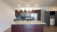 This 2-bedroom, 2-bathroom residence at The Zenith offers a thoughtfully designed single-floor layout that balances modern style with everyday functionality. The open-concept living and kitchen area serves as the heart of the unit, where rich dark cabinetry, a mosaic tile backsplash, and stainless steel appliances — including a gas range, built-in microwave, and French door refrigerator — come together to create a well-appointed culinary space. A large central island with a granite countertop and double sink is illuminated by a trio of elegant pendant lights, providing both additional prep space and a natural gathering point. Hardwood-style flooring flows seamlessly throughout the unit, lending warmth and continuity to every room. Each bedroom is generously proportioned, featuring ceiling fans, neutral wall tones, and windows that invite natural light. One bedroom opens directly to a private outdoor balcony via French doors. Both bathrooms are finished with granite-topped dark vanities and tiled tub-shower combinations. A dedicated in-unit laundry area adds convenience, while gray-paneled doors provide a contemporary accent throughout the space.