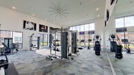 This well-appointed fitness center is designed to accommodate a wide range of workout routines within a single, open-plan space. The room is defined by soaring ceilings that create an airy, expansive atmosphere, complemented by large floor-to-ceiling windows along multiple walls that flood the interior with natural light and provide views of the surrounding outdoor greenery.

The flooring features a distinctive geometric-patterned carpet in shades of grey and blue, adding a modern, dynamic aesthetic throughout. Overhead, a large ceiling fan and recessed lighting ensure comfortable conditions during any workout session.

The fitness center is equipped with an extensive array of machines and equipment. Cardio offerings include treadmills, ellipticals, and a stair climber, while strength training is supported by a multi-functional cable machine, a dedicated punch bag station, kettlebells, a weight sled, and various resistance machines. Wall-mounted televisions are positioned throughout for entertainment. Large-scale athletic photography displayed on the walls adds a motivational, professional gym atmosphere. A mirrored wall further enhances the sense of space while supporting form-focused training.