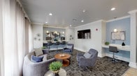 This single-floor communal lounge space is thoughtfully designed to serve both relaxation and productivity. The main lounge area is anchored by plush gray upholstered seating — including a curved sofa, swivel accent chairs, and a round wooden coffee table — arranged atop a patterned area rug that adds visual depth to the space. Recessed lighting and crown molding throughout lend the interior a polished, refined character.

A dedicated workstation nook, finished in a soft blue accent color, features a built-in desk with bar-height seating and pendant lighting, providing an ideal setting for focused work or study. A second compact desk station is also available nearby. A large wall-mounted flat-screen television is positioned prominently in the lounge, making the area well-suited for both casual gatherings and entertainment.

Natural light floods the space through a bank of large windows dressed with sheer curtains, creating a bright and welcoming atmosphere. The flooring transitions seamlessly from stone tile — visible in the entry corridor and surrounding hallways — to the carpeted lounge area. A dark credenza with decorative lamps and framed artwork completes the lounge's sophisticated aesthetic. The hallway connecting to additional areas is lined with wall sconces, maintaining a consistent and inviting ambiance throughout.