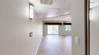 This thoughtfully designed studio in San Diego, CA (92127) offers a comfortable and functional single-floor layout with an open living area and two bathrooms. The main living space is finished with light wood laminate flooring that extends throughout, creating a warm and cohesive aesthetic. Recessed lighting illuminates the room, complemented by a textured ceiling and a mix of neutral and deep accent wall tones that add visual depth. A wall-mounted mini-split unit provides efficient climate control. Natural light pours in through a sliding glass door fitted with vertical blinds and decorative valances, leading to a private balcony with outdoor views of lush greenery. An additional window further brightens the space. The primary bathroom is a standout feature, boasting elegant marble-style tile flooring, a double-sink granite countertop vanity with framed mirrors, a soaking tub set on a raised platform, and a separate glass-enclosed shower stall — all illuminated by recessed ceiling lights. A second, more compact bathroom provides additional convenience with tile flooring. A hallway connects the living area to the bathrooms, completing this efficient and polished studio layout.