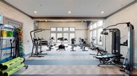 This fitness center is a thoughtfully designed, single-floor facility offering a comprehensive workout environment across several interconnected zones. The main cardio area is expansive and bright, featuring soaring ceilings with a striking wood-paneled upper section and large clerestory windows that flood the space with natural light. The room accommodates an extensive array of equipment, including treadmills, ellipticals, upright and recumbent bikes, stair climbers, and a rowing machine, all arranged to allow comfortable circulation.

A dedicated strength and functional training area houses a multi-station cable machine, free weights, and a dumbbell rack positioned alongside floor-to-ceiling mirrors that enhance the sense of space. A separate studio or spin room provides a more intimate setting, equipped with cycling bikes and a wall-mounted flat-screen TV recessed into wood-clad paneling, accompanied by a mirrored wall and a low platform with a handrail—suitable for stretching or barre-style workouts.

A transitional corridor connects the zones and includes a rack stocked with stability balls, medicine balls, and resistance accessories, along with a built-in bench and under-counter refrigeration. The flooring throughout combines light-toned patterned carpet tiles with contrasting blue striped sections, adding visual structure. Stone-accented accent walls contribute warmth and texture, while recessed lighting ensures consistent illumination throughout.