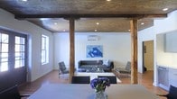 This distinctive single-floor space masterfully combines rustic and contemporary design elements to create a visually compelling environment. The most striking architectural feature is the richly toned, exposed wood plank ceiling supported by substantial raw timber posts, lending a warm industrial character that anchors the entire space. Recessed lighting is integrated throughout the ceiling, providing well-distributed ambient illumination that complements the natural light streaming in through the windows. The open layout flows seamlessly between a dining area and a living room, creating a cohesive and inviting atmosphere. The living area is furnished with a tufted leather sofa, modern accent chairs with chrome frames, and a glass coffee table set atop a plush area rug — all arranged around a bold abstract artwork that serves as a dynamic focal point on the wall. A wall-mounted split AC unit ensures year-round climate comfort. Hardwood flooring runs throughout, tying the spaces together with warmth and continuity. The dining area, positioned in the foreground, features a clean-lined table and sleek modern chairs, accented by a vibrant floral centerpiece. Together, these elements create a space that is both functional and aesthetically refined, ideal for entertaining or everyday living.