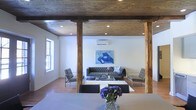 This distinctive single-floor space masterfully combines rustic and contemporary design elements to create a visually compelling environment. The most striking architectural feature is the richly toned, exposed wood plank ceiling supported by substantial raw timber posts, lending a warm industrial character that anchors the entire space. Recessed lighting is integrated throughout the ceiling, providing well-distributed ambient illumination that complements the natural light streaming in through the windows. The open layout flows seamlessly between a dining area and a living room, creating a cohesive and inviting atmosphere. The living area is furnished with a tufted leather sofa, modern accent chairs with chrome frames, and a glass coffee table set atop a plush area rug — all arranged around a bold abstract artwork that serves as a dynamic focal point on the wall. A wall-mounted split AC unit ensures year-round climate comfort. Hardwood flooring runs throughout, tying the spaces together with warmth and continuity. The dining area, positioned in the foreground, features a clean-lined table and sleek modern chairs, accented by a vibrant floral centerpiece. Together, these elements create a space that is both functional and aesthetically refined, ideal for entertaining or everyday living.