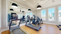 Located at 2670 Meadow Creek Rd in Kissimmee, Florida, this fitness center is a thoughtfully designed, single-floor facility built to support a comprehensive workout experience. The space features light-toned hardwood-style flooring throughout, neutral gray walls, and crisp white trim, creating a clean and energizing atmosphere. Large windows line multiple walls, flooding the interior with abundant natural light and offering views of the surrounding outdoor amenity areas, including a glimpse of a pool and lush tropical landscaping. Floor-to-ceiling mirrors are strategically placed to enhance the sense of space and assist with form during exercise. The cardio zone is well-appointed with multiple treadmills and elliptical cross-trainers, while the free weight area includes a full rack of dumbbells ranging in various weights alongside adjustable workout benches. A cable machine and additional strength equipment round out the offerings for a balanced training environment. Overhead, bold black pendant lights paired with a ceiling fan provide both stylish illumination and comfortable airflow. A wall-mounted television and posted fitness center guidelines add to the facility's functional, user-friendly design. Double glass doors open directly to the outdoor amenity area, further connecting the interior to the sunny Florida surroundings.