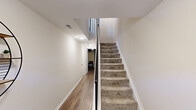 This three-story residence in Novi, Michigan offers a well-organized layout distributed across a ground level, main floor, and upper floor. The entry-level floor provides a utility room and hallway, finished with durable wood-look flooring and carpeted stairs with a dark metal railing connecting the levels — offering practical space for storage and everyday function.

The main floor, the largest of the three levels, serves as the social hub of the home. A combined kitchen and living room creates an open-concept atmosphere ideal for both daily living and entertaining. The kitchen is outfitted with white shaker-style cabinetry, light countertops, stainless steel appliances including a refrigerator, range, and over-the-range microwave, and a classic white subway tile backsplash over wood-look flooring. The living area features neutral tones and natural light from surrounding windows.

The upper floor houses both bedrooms, a dedicated office, and two full bathrooms. The primary bedroom is generously sized with carpet flooring, natural light from a window, and a closet. The second bedroom offers similar comfort, while the office provides a windowed, carpeted space suitable for remote work. One bathroom features a walk-in shower with a frosted sliding glass door. A spacious walk-in closet with wire shelving adds practical storage.