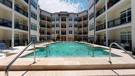 This amenity space offers two distinct areas designed to support both relaxation and active fitness. The outdoor pool area serves as a central courtyard feature, framed by a multi-story building with balconies on all sides. The generously sized rectangular pool features clean tile coping, built-in entry railings, and decorative water jets. The surrounding deck is finished in light-toned pavers and furnished with lounge chairs and shaded umbrella seating, creating a resort-like atmosphere open to the sky.

The fitness center is a well-appointed, multi-zone space finished with patterned herringbone-style carpet flooring throughout. Soaring ceilings and an expansive wall of mirrors visually enlarge the space while ceiling fans maintain airflow. Large windows along multiple walls bring in abundant natural light and offer outdoor views. The strength training zone is comprehensively stocked with a cable multi-rack system, power rack, adjustable bench, and a full dumbbell rack ranging across multiple weights. Functional training equipment includes medicine balls, battle ropes, and stability balls. Cardio equipment includes treadmills, a Precor recumbent bike, and a WaterRower rowing machine. The layout provides ample open floor space for stretching or bodyweight exercises.