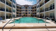 This amenity space offers two distinct areas designed to support both relaxation and active fitness. The outdoor pool area serves as a central courtyard feature, framed by a multi-story building with balconies on all sides. The generously sized rectangular pool features clean tile coping, built-in entry railings, and decorative water jets. The surrounding deck is finished in light-toned pavers and furnished with lounge chairs and shaded umbrella seating, creating a resort-like atmosphere open to the sky.

The fitness center is a well-appointed, multi-zone space finished with patterned herringbone-style carpet flooring throughout. Soaring ceilings and an expansive wall of mirrors visually enlarge the space while ceiling fans maintain airflow. Large windows along multiple walls bring in abundant natural light and offer outdoor views. The strength training zone is comprehensively stocked with a cable multi-rack system, power rack, adjustable bench, and a full dumbbell rack ranging across multiple weights. Functional training equipment includes medicine balls, battle ropes, and stability balls. Cardio equipment includes treadmills, a Precor recumbent bike, and a WaterRower rowing machine. The layout provides ample open floor space for stretching or bodyweight exercises.