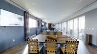 The Sky Lounge is a refined and contemporary elevated gathering space designed to impress. The open-plan layout is anchored by a striking large natural wood dining table surrounded by distinctive woven-strap chairs, creating a dramatic focal point that blends organic materials with modern design sensibilities. The space is bathed in abundant natural light, courtesy of floor-to-ceiling windows and sliding glass panels that line the perimeter, offering sweeping panoramic views of the surrounding skyline and cityscape. Deep slate-blue accent walls, complemented by elegant floor-length drape panels, lend the interior a rich, sophisticated atmosphere. Recessed lighting throughout the smooth white ceiling ensures the space remains well-lit at all times of day. Toward the rear, a curated arrangement of contemporary artwork — including an abstract sculptural piece — adds a gallery-like quality to the lounge. The hardwood flooring grounds the space with warmth, providing a refined contrast to the cool, modern palette. Whether used for private dining, corporate events, or social gatherings, the Sky Lounge delivers an atmosphere of elevated luxury and thoughtful design, making it a versatile and memorable destination for any occasion.