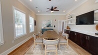 Located at 12 Wilkinson Way in Bluffton, SC 29910, this single-floor space functions as a well-appointed meeting or conference room designed for both comfort and functionality. The room centers around a large rectangular herringbone-patterned wood table surrounded by upholstered arm chairs with nailhead trim detailing, all set atop a neutral area rug over warm hardwood flooring. Crown molding and recessed lighting run throughout the ceiling, complemented by ceiling fans that enhance air circulation and comfort. One wall features a built-in dark espresso cabinetry unit with a light marble-style countertop, above which floating white shelves display decorative accents and a wall-mounted flat-screen television — ideal for presentations or group viewings. A teal-painted sideboard adds a pop of color alongside a large abstract artwork, contributing to the room's coastal-inspired aesthetic. Multiple double-hung windows fitted with white wood blinds line the walls, flooding the space with natural light and offering views of the surrounding neighborhood. The soft, neutral warm-beige and muted blue-gray wall tones create a calm, professional atmosphere well-suited for meetings, collaborative gatherings, or community events.