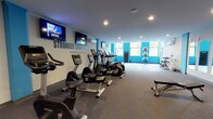 Located at 1401 N 5th St in Philadelphia's vibrant 19122 neighborhood, this fitness center offers a comprehensive single-floor workout facility designed to accommodate a wide range of exercise needs. The space is defined by a clean, modern aesthetic featuring a neutral gray and bold blue color scheme, with motivational phrases such as "Motivation Determines What You Do" and "Stronger Than Yesterday" displayed prominently across the walls to inspire members during their workouts.

The cardio zone is well-equipped with multiple treadmills, upright and recumbent stationary bikes, and elliptical machines, all arranged to allow comfortable spacing between users. Wall-mounted flat-screen TVs are positioned throughout for entertainment during workouts. A dedicated strength training area includes a cable and pulley rack system, a rowing machine, and a heavy punching bag, catering to both functional and resistance training. The free weight section features a full dumbbell rack alongside open floor space suitable for stretching or floor exercises.

Large windows along one wall invite ample natural light and outdoor views, enhancing the overall atmosphere of the space. Full-length mirrors and a water station add practical amenities. The flooring transitions between carpet and rubberized mat surfaces, providing appropriate traction and cushioning throughout the facility. Recessed lighting and a wall-mounted HVAC unit ensure a comfortable environment year-round.