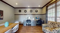 This business center in Memphis, Tennessee, is a thoughtfully designed single-floor workspace that balances productivity with comfort. The room features warm hardwood flooring throughout, complemented by two-tone walls with classic beadboard wainscoting trimmed in rich dark wood molding that extends along the ceiling line, lending the space a polished, traditional character.

A built-in workstation along one wall accommodates two desktop computers side by side, each positioned at a granite-topped desk with gray cabinetry below for storage. A printer is also available, making the station fully equipped for everyday business tasks. Four decorative sunburst-style wall medallions are evenly spaced above the workstation, adding a refined aesthetic touch.

A rustic natural wood conference table with white upholstered chairs occupies the center of the room, offering a comfortable collaborative area for meetings or group work. A patterned accent chair provides additional informal seating near a large window dressed with wooden blinds, allowing natural light to filter in while maintaining privacy.

A bold abstract canvas artwork decorates one wall, adding visual interest. French doors with glass panels lead to an outdoor area, further brightening the space and providing easy access to the exterior. Recessed ceiling lighting ensures the room remains well-lit throughout.