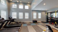 Located at 4964 NC-55 Hwy in Cary, North Carolina, this fitness center offers a thoughtfully designed, single-floor workout facility built to accommodate a wide range of exercise needs. The space is defined by dramatically tall ceilings that create an open, airy atmosphere, complemented by an abundance of natural light streaming through multiple large windows arranged in stacked rows along the walls. Soft blue-gray walls and patterned modular carpet tiles in neutral tones of beige, gray, and blue contribute to a clean, modern aesthetic throughout.

The main workout area is generously proportioned, housing a well-rounded selection of cardio equipment including treadmills, a rowing machine, a stationary spin bike, and an elliptical trainer. A Precor functional training system provides strength and resistance training options, while plyo boxes and balance tools support functional fitness routines. A dedicated stretch and open-floor zone offers additional versatility for yoga or bodyweight exercises.

A secondary area features built-in cabinetry, a compact refreshment station, and motivational wall graphics, serving as a convenient wellness hub within the facility. Ceiling fans, recessed lighting, and sanitization stations are thoughtfully integrated throughout, ensuring comfort and cleanliness. The overall design balances functionality with an inviting, light-filled ambiance ideal for an effective workout experience.