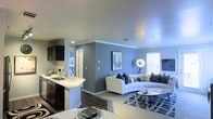This single-floor space showcases an open-concept layout that integrates the kitchen, dining, and living areas into one cohesive environment. The kitchen is outfitted with dark cabinetry, a white countertop peninsula with an undermount sink, and modern fixtures, creating a striking contrast that adds visual depth. The peninsula doubles as a casual dining or entertaining surface, enhancing the space's functionality.

The living area features a bold blue accent wall that serves as a focal point, complemented by neutral grey tones throughout. Crown molding adds a refined architectural detail along the ceiling. Plush carpeting in the living zone transitions from the hard flooring in the kitchen, delineating the two areas while maintaining a unified aesthetic.

Natural light enters through a window fitted with blinds, brightening the space alongside recessed lighting and a floor lamp. The living area comfortably accommodates a sectional sofa and armchair arrangement centered around a round coffee table, all styled over a patterned area rug. A hallway visible in the background suggests additional private rooms beyond the main living space.