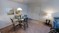This single-floor residence offers an open and functional layout centered around a combined dining and living area. The dining space is highlighted by a round glass-top table paired with four black metal chairs featuring cushioned seats, positioned beneath a classic white pendant light that provides focused illumination. Soft, neutral-toned wall-to-wall carpeting flows throughout the space, lending a comfortable and cohesive feel. The walls are painted in a light, muted tone, creating a calm and airy ambiance. A framed floral artwork adds a subtle decorative touch to the dining area. The entryway, located at the far end of the room, features a paneled white front door with a small area rug placed at the threshold. An additional interior door is also visible, likely leading to a closet or adjoining room. The overall layout is straightforward and practical, with ample open floor space between the dining area and the entryway, allowing for easy movement throughout. The design prioritizes simplicity and functionality, making the space well-suited for everyday living.