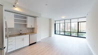 This single-floor unit showcases a contemporary design characterized by a bright, open-concept living and kitchen area. The living space is anchored by a dramatic floor-to-ceiling window wall framed in dark steel, allowing abundant natural light to fill the interior while offering elevated views of the surrounding urban skyline. Light-toned hardwood flooring runs throughout the main living areas, contributing to an airy, cohesive aesthetic.

The kitchen is well-appointed with white cabinetry, granite countertops, a subway tile backsplash, and a full suite of stainless steel appliances including a refrigerator, range, microwave, and dishwasher. Track lighting overhead enhances functionality and adds a modern touch. Open shelving provides additional display and storage options.

A hallway with a sliding closet door connects the main living area to the private rooms. The bedrooms feature neutral carpeting and windows with city views, offering a calm retreat. The bathroom includes a soaking tub and shower combination with white subway tile accented by a decorative mosaic border, white cabinetry, and granite countertops, maintaining the unit's clean, modern character throughout.