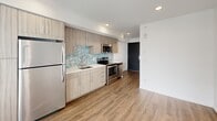 Located on the fourth floor at 5512 17th Ave NW in Seattle's Ballard neighborhood, Unit 411 is a thoughtfully designed studio that maximizes every inch of its compact footprint. The open-plan kitchen and living area flow seamlessly together, anchored by warm wood-tone luxury vinyl plank flooring throughout. The kitchen is outfitted with contemporary flat-front cabinetry in a light wood finish, stainless steel appliances including a refrigerator, electric range, and over-the-range microwave, a white quartz countertop, and an eye-catching teal and neutral mosaic tile backsplash that adds a pop of color and texture. Recessed lighting keeps the space bright and modern. The living area is clean and open, with crisp white walls and a large floor-to-ceiling window with blinds that floods the room with natural light and provides access to a private patio, extending the living space outdoors. The bathroom continues the mosaic tile motif, featuring a full accent wall of blue and beige mosaic tiles framing the mirror and vanity, complemented by a floating wood-tone vanity with a white countertop, tile flooring, and a full soaking tub. A bold dark sliding barn-style door adds a striking design contrast at the entry, completing this stylish and efficient urban residence.