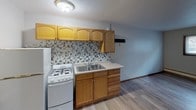 This single-floor studio is a well-organized space designed to maximize functionality within a compact footprint. The main living area features light gray wood-look vinyl flooring that runs throughout, providing a cohesive and easy-to-maintain surface. A large window with horizontal blinds allows natural light to fill the room, while a wall-mounted air conditioning unit and baseboard heating ensure year-round comfort.

The kitchen is efficiently laid out along one wall and includes warm honey-toned wood cabinetry, both upper and lower, paired with a mosaic tile backsplash in neutral gray and beige tones. The kitchen is equipped with a white gas range, refrigerator, and a double-basin sink with ample counter space for meal preparation.

A mirrored sliding closet provides useful storage and visually expands the living area, reflecting light across the room. The entry features a solid wood door, adding a classic touch. The bathroom is fitted with a tub and shower combination, with full-height beige ceramic tile surround offering a clean and durable finish. The textured ceiling throughout adds a subtle architectural detail to the overall design.