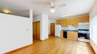 This single-floor unit offers a practical and well-organized layout, combining a kitchen and living area within one open space, complemented by a full bathroom. Light wood laminate flooring runs throughout, giving the space a clean and cohesive look.

The kitchen is equipped with warm honey-toned wooden cabinetry extending to the ceiling for ample storage, a white refrigerator, an electric range with overhead ventilation, a black dishwasher, and a single-basin sink with a window nearby. Fluorescent ceiling lighting ensures the kitchen area is well-lit at all times.

The living area benefits from a large picture window fitted with vertical blinds, allowing generous natural light to pour in while framing views of surrounding greenery. A ceiling fan overhead provides year-round comfort. The open layout connects the living and kitchen zones seamlessly, creating a functional and airy feel despite the compact footprint. A sliding door closet and an entry door are also visible from the main area, offering practical storage and access.

The bathroom features light wood flooring continuing from the main area, a wall-mounted vanity with a distinctive blue countertop and integrated sink, a large mirror above, and a full-size shower-tub combination. The overall aesthetic of the unit is clean, minimal, and functional, making efficient use of every square foot.