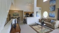This 3-floor residence makes an immediate impression with its dramatic vaulted ceilings that rise well above the main living area, creating an expansive, airy atmosphere throughout the open-concept floor plan. The living room serves as the central hub, featuring a glass-top coffee table, plush seating, and a decorative area rug that anchors the space. A round porthole-style window on the upper wall adds an architectural focal point while drawing in additional natural light. The kitchen is positioned at the heart of the home, featuring light wood cabinetry, dark countertops, and black appliances including a refrigerator and microwave. A raised breakfast bar with barstool seating provides a casual dining option and creates a natural divide between the kitchen and the main living space. An adjacent dining area accommodates a full table and chair set, ideal for more formal meals. Track lighting and a chandelier provide warm ambient illumination throughout. The multi-level layout across three floors offers distinct zones for living, sleeping, and additional functionality, making this home both versatile and well-suited for a variety of lifestyles.