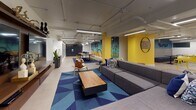 This single-floor amenity space is a dynamic, thoughtfully designed common area that blends comfort, functionality, and bold aesthetics. The layout follows an open, L-shaped floor plan that seamlessly connects multiple distinct zones, encouraging both socializing and individual use.

The central lounge area anchors the space with a generously sized sectional sofa in charcoal grey, arranged atop a striking geometric blue area rug. A wooden coffee table and a mid-century modern accent chair complete the seating arrangement, while an abstract canvas painting adorns the wall above, adding a creative focal point. Bright yellow accent walls punctuate the space throughout, creating an energetic contrast against the white and neutral tones.

Adjacent to the lounge, a communal co-working zone features a live-edge wood table with industrial trestle legs, surrounded by yellow metal stools — an inviting spot for collaborative work or casual dining. A wall of individual mailboxes lines one side, offering practical resident services seamlessly integrated into the design.

The game area introduces a bold yellow foosball table alongside cozy booth-style seating with patterned tile accents, perfect for recreation and informal gatherings. Exposed ceiling infrastructure with linear pendant and track lighting lends an industrial edge, while polished concrete flooring ties the entire space together. The result is a lively, welcoming environment with a distinctly urban character.