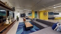 This single-floor amenity space is a dynamic, thoughtfully designed common area that blends comfort, functionality, and bold aesthetics. The layout follows an open, L-shaped floor plan that seamlessly connects multiple distinct zones, encouraging both socializing and individual use.

The central lounge area anchors the space with a generously sized sectional sofa in charcoal grey, arranged atop a striking geometric blue area rug. A wooden coffee table and a mid-century modern accent chair complete the seating arrangement, while an abstract canvas painting adorns the wall above, adding a creative focal point. Bright yellow accent walls punctuate the space throughout, creating an energetic contrast against the white and neutral tones.

Adjacent to the lounge, a communal co-working zone features a live-edge wood table with industrial trestle legs, surrounded by yellow metal stools — an inviting spot for collaborative work or casual dining. A wall of individual mailboxes lines one side, offering practical resident services seamlessly integrated into the design.

The game area introduces a bold yellow foosball table alongside cozy booth-style seating with patterned tile accents, perfect for recreation and informal gatherings. Exposed ceiling infrastructure with linear pendant and track lighting lends an industrial edge, while polished concrete flooring ties the entire space together. The result is a lively, welcoming environment with a distinctly urban character.