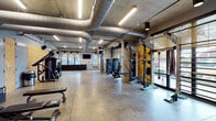 This fitness facility presents a distinctly industrial aesthetic, characterized by exposed concrete ceilings, large silver HVAC ductwork running overhead, and polished concrete floors throughout. The open layout provides generous floor space, allowing equipment to be arranged with ample room between stations for comfortable use.

The main workout floor is well-lit by linear LED fixtures suspended from the ceiling and benefits from abundant natural light through large windows and glass entry doors that line the perimeter walls, offering views of the surrounding exterior. Strength training equipment, including cable machines, power racks with wooden accent panels, selectorized machines, and flat benches, is distributed throughout the main floor.

A dedicated free weights room features organized racks of dumbbells, medicine balls, weight plates, and a flat utility bench along textured block walls, providing a focused environment for strength training.

Near the back of the facility, a reception or check-in counter area is finished with dark cabinetry, pendant lighting, built-in shelving, and large windows dressed with roman shades, adding a polished, functional touch. A wall-mounted television is also present for entertainment. The overall ambiance is clean, open, and purpose-driven.