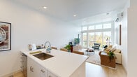 Located at 7007 SW Hampton St in Tigard, Oregon, this thoughtfully designed one-bedroom, one-bathroom unit offers a clean, modern aesthetic across its efficiently arranged single floor. The open-concept living and kitchen area serves as the heart of the home, where a galley-style kitchen features white cabinetry with brushed hardware, sleek quartz countertops, stainless steel appliances including a built-in microwave and range, and an undermount sink with a contemporary faucet. The kitchen flows seamlessly into the living room, which is anchored by striking floor-to-ceiling windows that flood the space with natural light and frame scenic views of the surrounding Tigard treeline. Light wood-tone flooring runs consistently throughout the unit, enhancing the bright, airy atmosphere. The bedroom is a comfortable retreat, offering a well-proportioned layout with a closet and the same warm flooring. The full bathroom is finished with white cabinetry, a single vanity with an integrated sink, and a tub-shower combination with clean white tile surround. Recessed lighting throughout and crisp white walls complete the contemporary look, making this a well-appointed and efficiently designed urban residence.