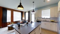 This single-floor space showcases a thoughtfully designed open-concept layout that integrates the kitchen, dining, and living areas into one cohesive environment. The kitchen serves as the focal point, anchored by a generously sized island with a sleek dark countertop, offering both additional prep space and casual dining seating. Two industrial-style black pendant lights hang above the island, adding a contemporary aesthetic touch. Cabinetry is finished in a clean off-white tone with brushed silver hardware, complementing the light wood flooring that runs throughout. A stainless steel sink is positioned beneath a window near the perimeter counter, which also features a white subway tile backsplash. The adjoining living and dining area is bathed in natural light from multiple large windows dressed with white blinds and dark curtain panels. Accent walls in warm terra cotta and soft blue tones introduce color and visual depth to the space. The living area is furnished with comfortable seating, creating a welcoming and functional atmosphere. Overall, the open layout promotes a sense of spaciousness and easy flow between the key living zones.