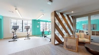 The Fitness Center Upper Level is a thoughtfully designed workout facility that combines functional fitness spaces with contemporary aesthetics. The space is defined by bold teal-painted walls that energize the environment, contrasting with white ceilings and neutral flooring throughout.

The exercise area is well-equipped with cardio machines including stationary bikes and rowing machines, all positioned to take advantage of the large, multi-pane windows that flood the room with natural light and offer views of the surrounding exterior. A wall-mounted mirror expands the sense of space within the workout zone.

A standout architectural feature is the dramatic diagonal wood-and-glass partition that elegantly divides the fitness floor from an adjacent lounge area. This sculptural element incorporates warm walnut tones with geometric open shelving, adding visual depth and sophistication. The lounge area features woven rattan chairs and a low round coffee table, providing a comfortable place to rest before or after a workout.

A separate entry lounge area includes carpeted flooring, a wall-mounted flat-screen television set against a teal accent wall, built-in cabinetry, and glass-paneled doors leading to the main fitness floor. Recessed lighting throughout ensures even, welcoming illumination across all zones of the facility.